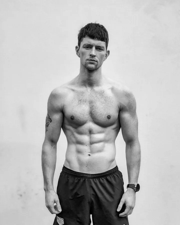 Homme européen aux cheveux bruns-noirs, torse nu, posant près d'une échelle peinte, regard intense, en intérieur sombre.