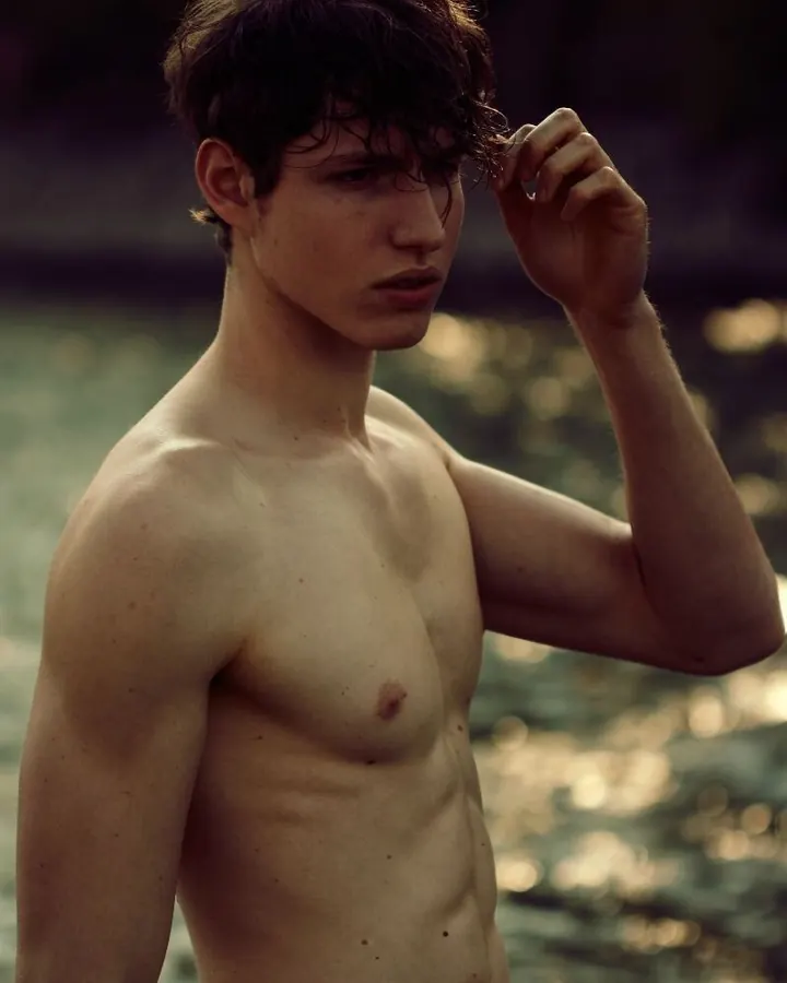 Jeune homme européen aux cheveux châtains, torse nu, debout près de l'eau, la main dans les cheveux humides, regard intense.