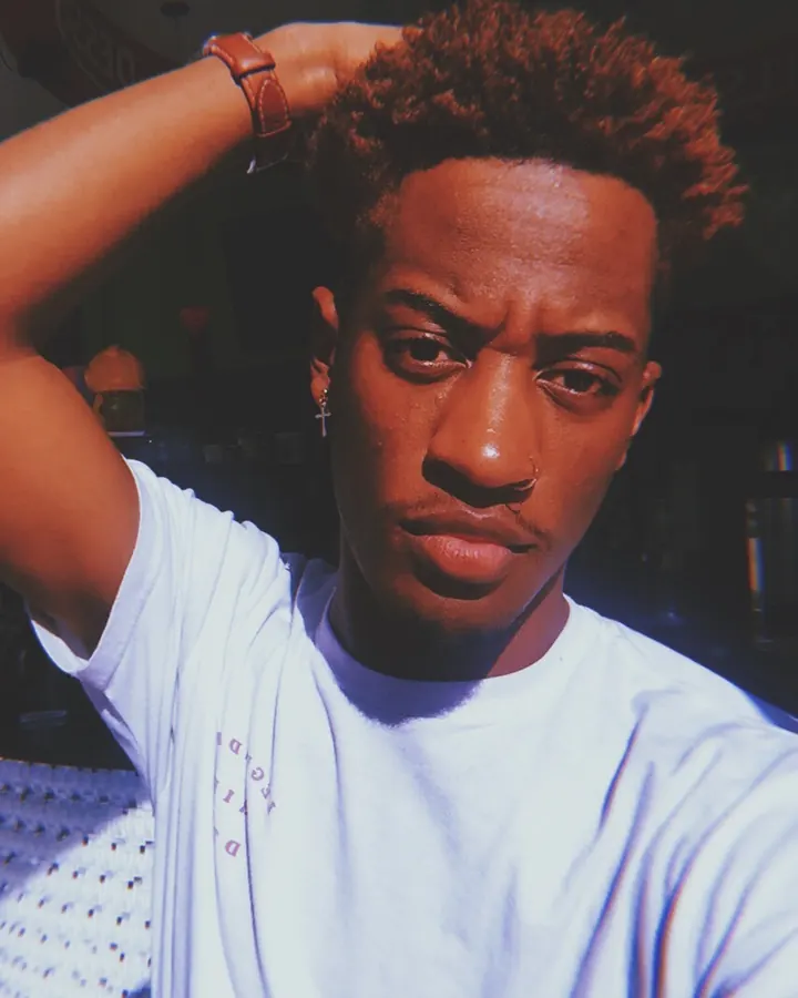 Homme afro aux cheveux bruns-noirs, portant un t-shirt blanc, la main dans les cheveux, regard intense sous une lumière vive.