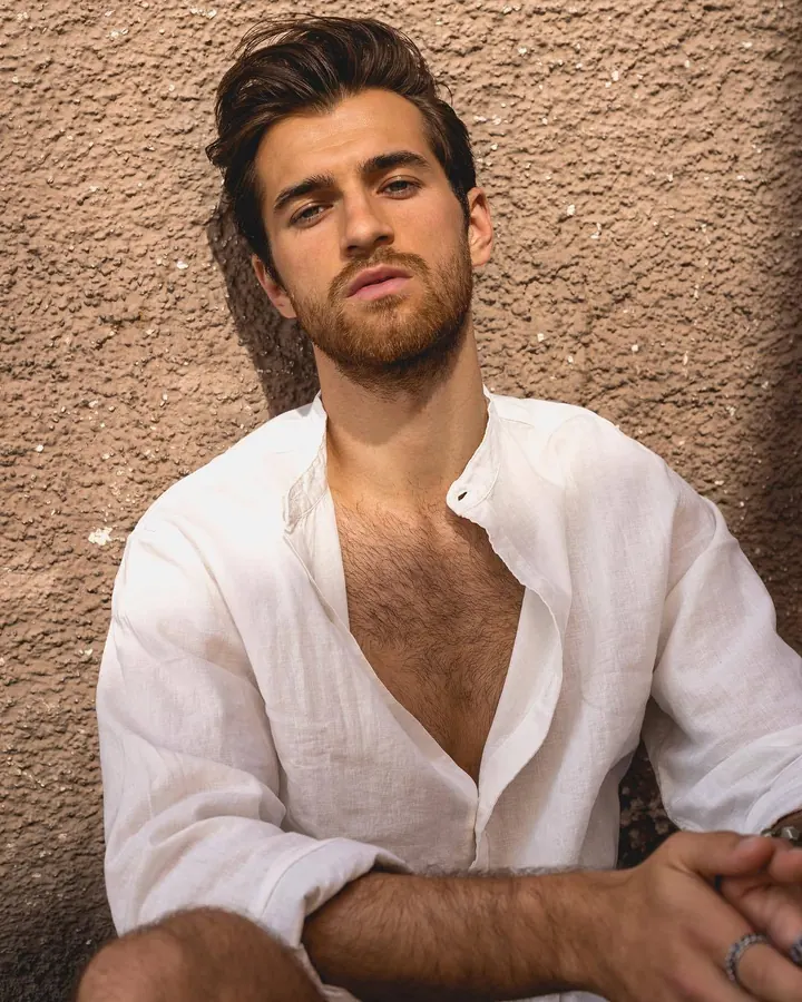 Homme européen aux cheveux châtains, torse nu, regardant l'appareil photo avec un air sérieux, en extérieur contre un mur beige.