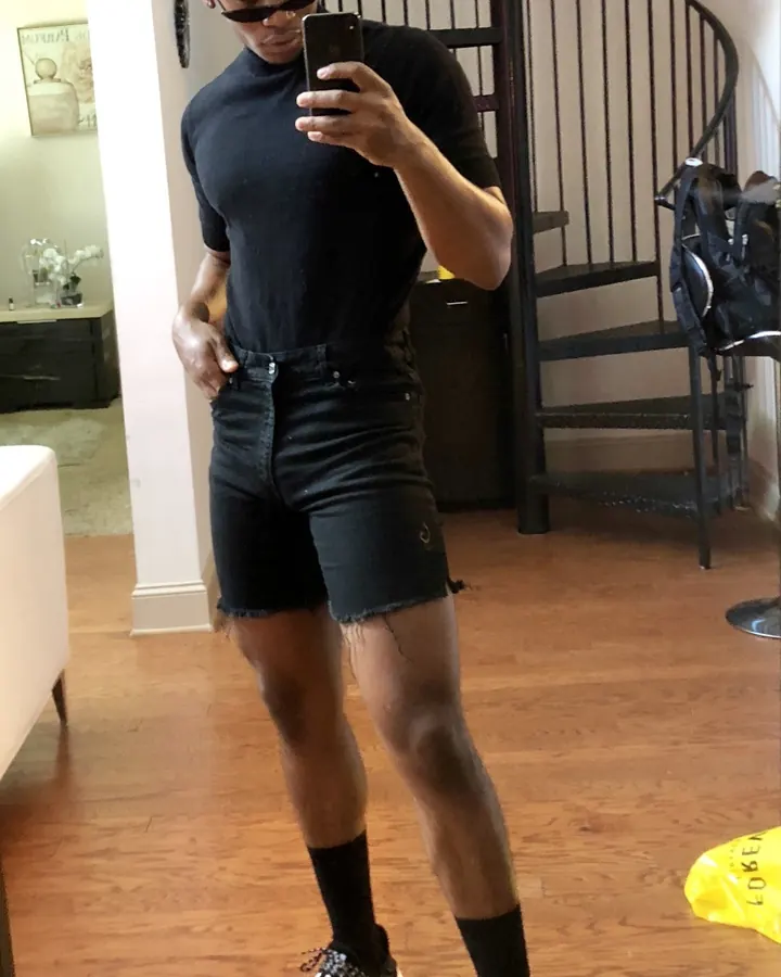 Homme afro avec cheveux blonds, lunettes de soleil, t-shirt noir et shorts noirs, prenant un selfie devant un miroir dans un intérieur moderne.
