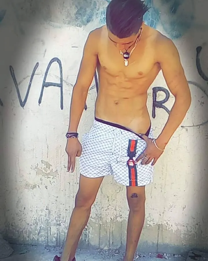 Homme indien aux cheveux bruns-noirs, torse nu, posant devant un mur graffiti, mains sur les hanches, regard vers le bas.