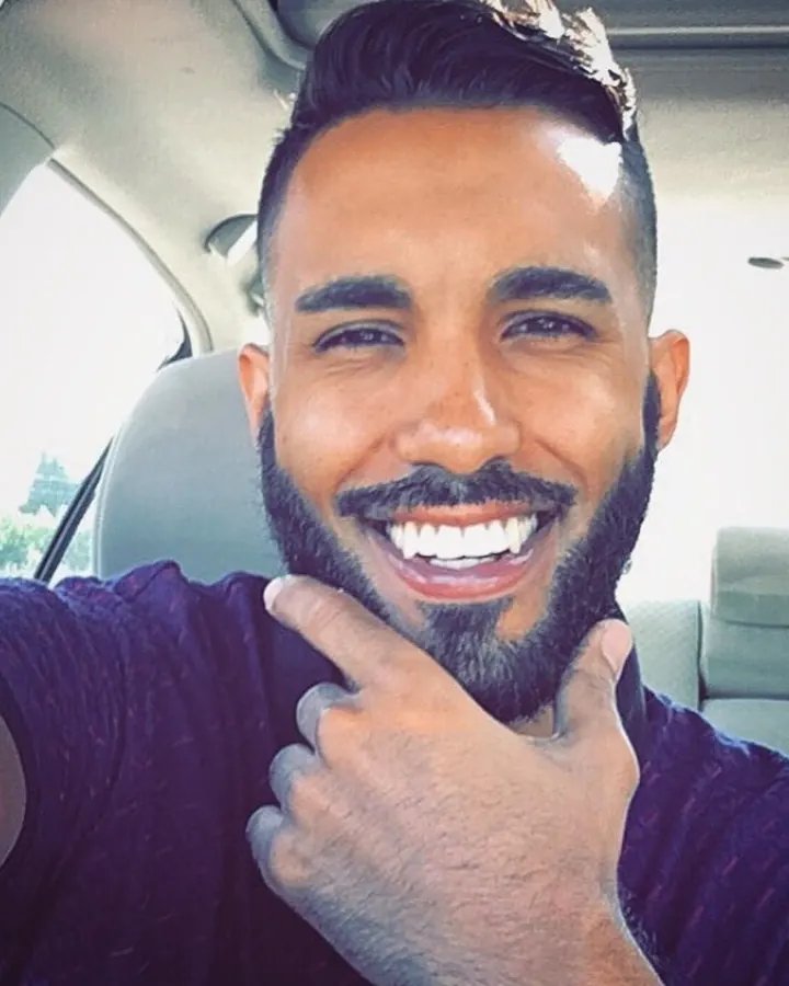 Homme souriant, barbu, avec des cheveux foncés coiffés en arrière, portant un t-shirt violet, assis dans une voiture.
