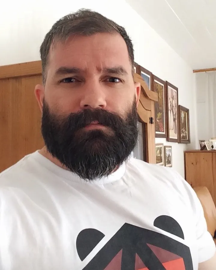 Homme barbu, regardant l'objectif, portant un t-shirt blanc avec un logo géométrique noir et rouge.