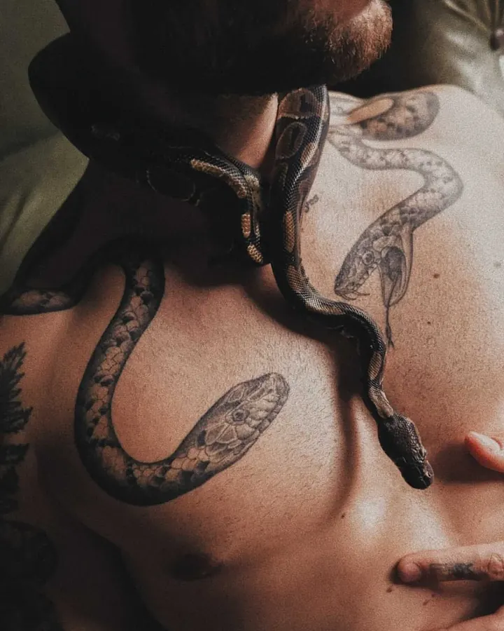 Plan rapproché d'un homme barbu, torse nu, avec un serpent réel enroulé autour de son cou et de sa poitrine tatouée.