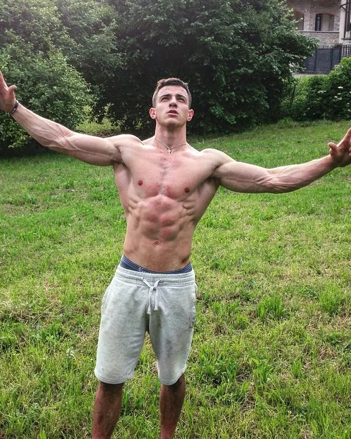 Homme torse nu, les bras écartés, debout dans un jardin, portant un short gris clair, avec une croix en collier.