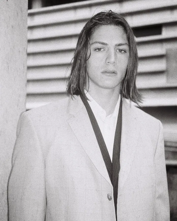 Portrait en noir et blanc d'un jeune homme aux cheveux mi-longs, portant un costume clair et une chemise blanche, regardant l'objectif.