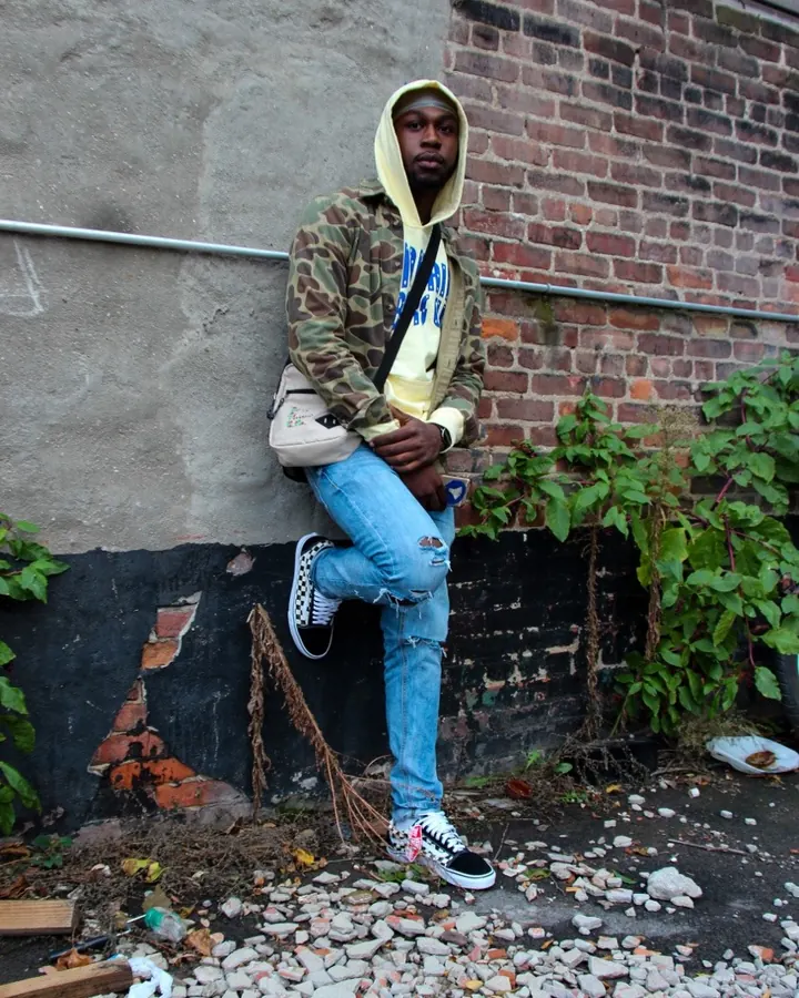 Homme assis contre un mur de briques, portant une veste camouflage, un sweat jaune à capuche, un sac bandoulière, et des baskets Vans.