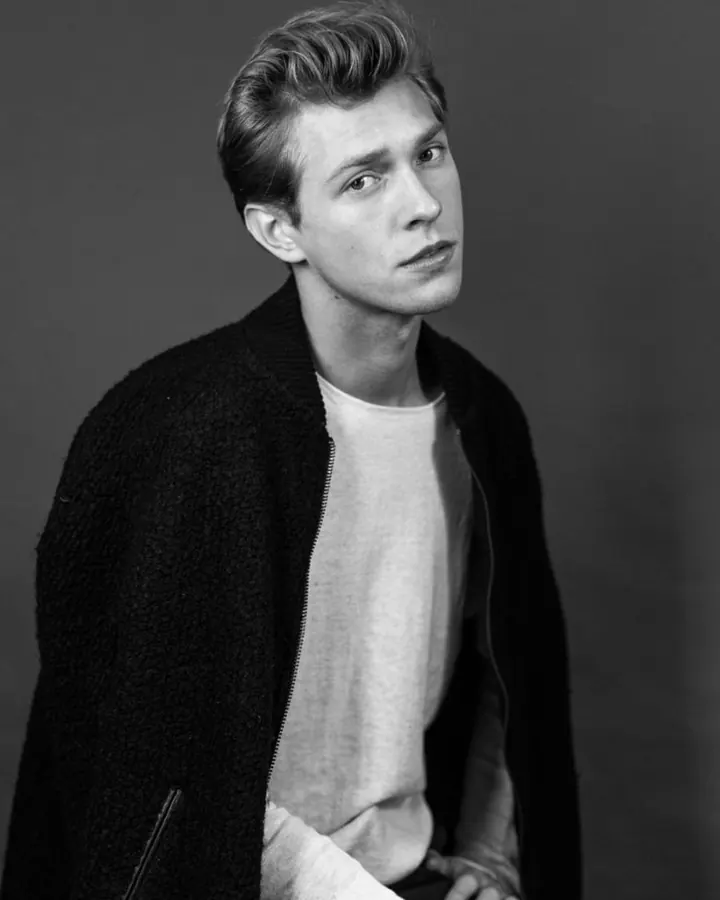 Homme en noir et blanc, cheveux blonds coiffés en arrière, vêtu d'une veste noire et d'un t-shirt blanc, regardant vers le haut, assis sur une chaise