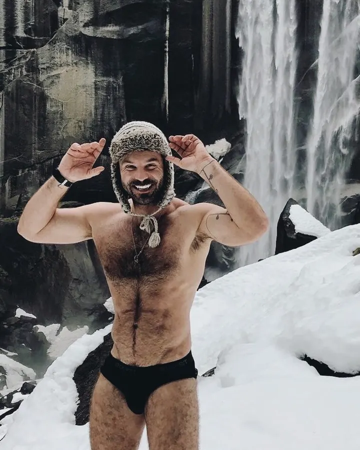 Homme torse nu, souriant, portant un bonnet en fourrure, debout dans la neige devant une chute d'eau gelée, tenant le bonnet avec les mains