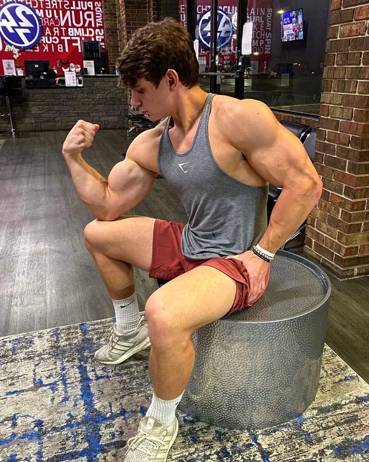 Jeune homme aux cheveux bruns bouclés, vêtu d'un débardeur gris et de shorts rouges, assis sur un tabouret, faisant un geste de flexion du biceps dans une salle de sport
